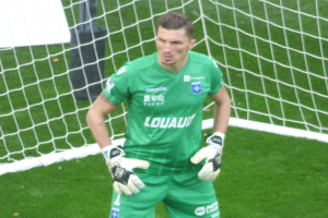 Ein Torwart in grüner Uniform steht vor einem Fußballtor auf saftig grünem Rasen, Handschuhe tragend.