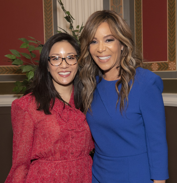Zwei Frauen stehen nebeneinander und lächeln auf der Women in Film Pre-Oscar-Luncheon 2019, eine in einem blauen Kleid und die andere in einem roten Kleid mit Brille, vor einer Pflanze und einem Vorhang.