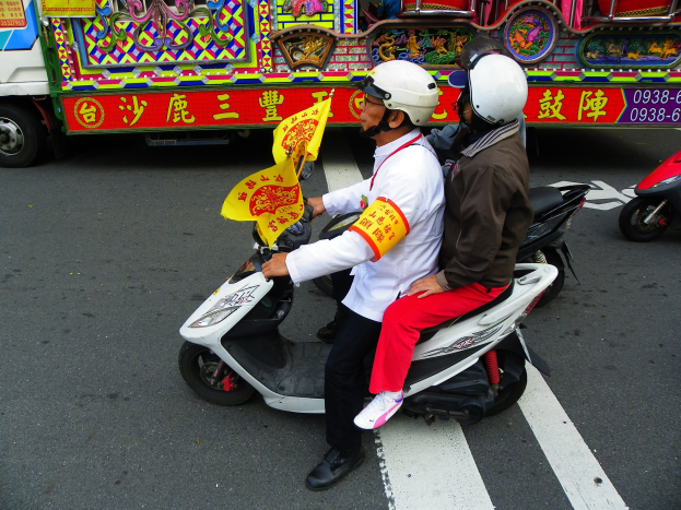 Zwei Menschen mit Helmen fahren auf einem Motorroller, einer hält eine gelbe Fahne, mit einem Fahrzeug im Hintergrund.