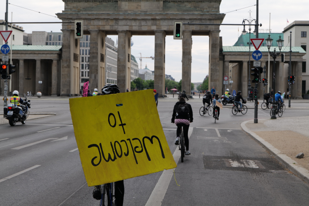 Eine Gruppe von Radfahrern fährt am Brandenburger Tor in Berlin vorbei, einer hält ein gelbes Schild, mit Laternenmasten, Verkehrszeichen, Gebäuden, Bäumen und einem klaren Himmel im Hintergrund.