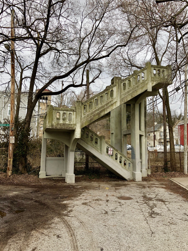 Eine alte Brücke überspannt einen Schotterweg in einer Stadt, umgeben von Bäumen, Pfählen und Gebäuden, mit dem Himmel im Hintergrund.
