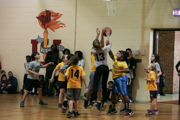 Kinder in gelben und grauen T-Shirts spielen Basketball, ein Mädchen in Grau mit der Nummer 13 wirft den Ball, ein Mann in Blau sitzt links, eine weiße Wand mit Text dahinter und ein Ausgangsschild sowie ein rotes Backsteingebäude rechts.