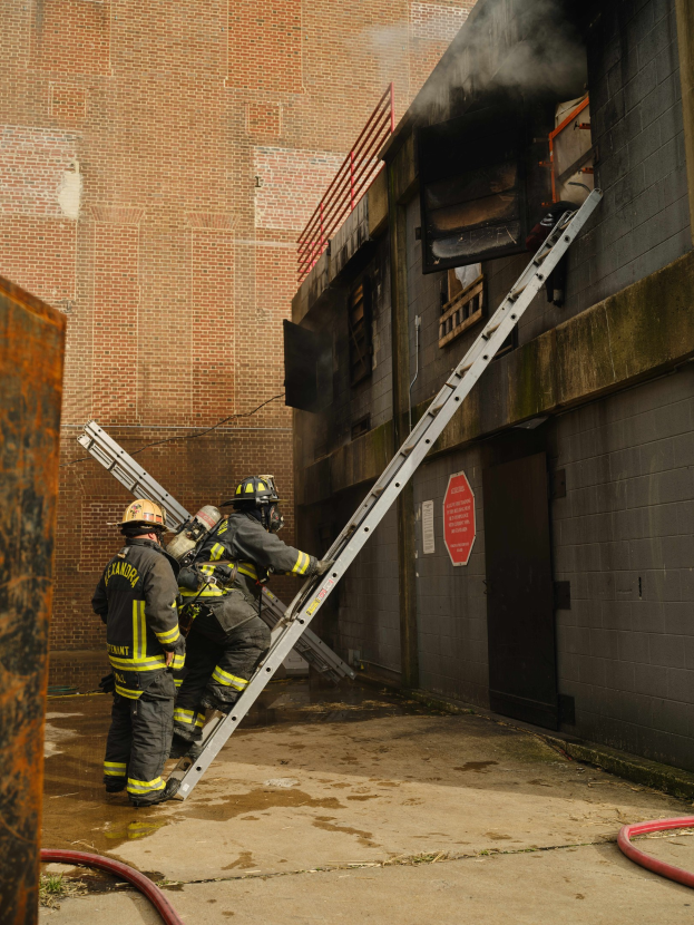 Feuerwehrleute mit Helmen klettern an einer Leiter zu einem raucherfüllten Gebäude hoch, mit einem Metallobjekt links und Rohren auf dem Boden.