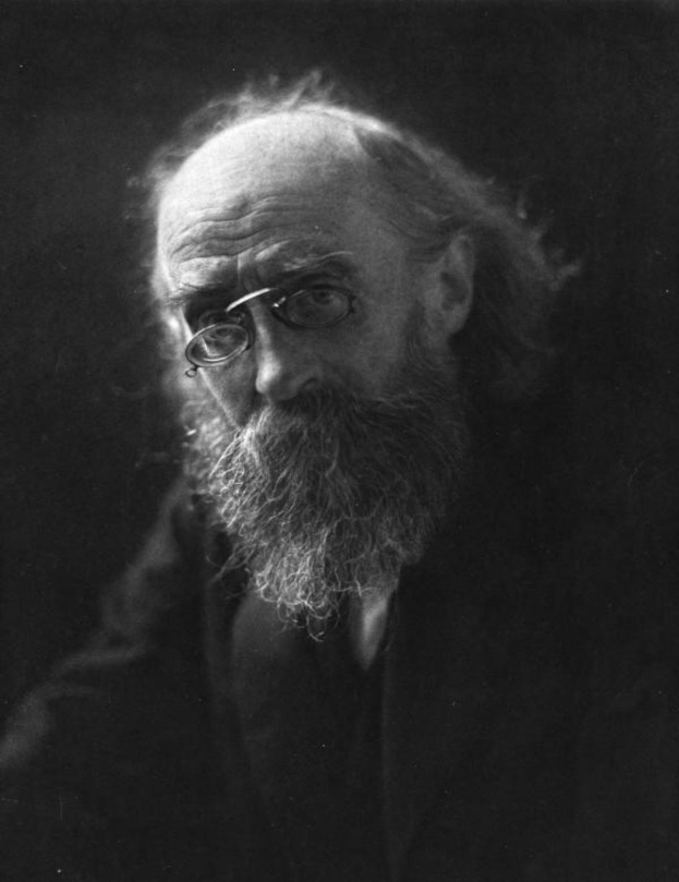 Schwarz-weiß-Porträt eines alten Mannes mit langem Bart und Brille vor dunklem Hintergrund.