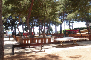 Ein Park mit einem zentralen Tischtennis-Tisch, umgeben von Bänken, Schaukeln, Bäumen und anderen Gegenständen, mit Blick auf einen See unter einem klaren blauen Himmel.