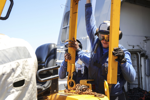Zwei Männer in blauen Uniformen und Schutzausrüstung arbeiten an einem gelben Gabelstapler, wobei einer ein Metallobjekt hält, während im Hintergrund ein Fahrzeug und der Himmel zu sehen sind.