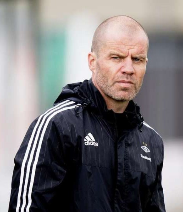 Baldiger Trainer in marineblauer Jacke mit weißen Streifen und weißem Kragen auf einem Fußballfeld mit gefalteten Händen und ernster Miene.