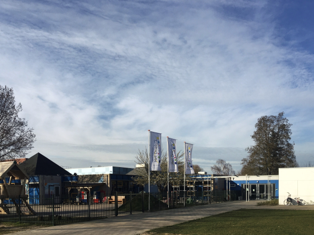 Gro├če Geb├Ąude mit Metallzaun, Rasenfl├Ąche, Weg, B├Ąumen, Pfosten, Schildern, einem abgestellten Fahrrad, n├Ąheren H├Ąusern und einem bew├Âlktem Himmel, identifiziert als neuer Spielplatz an der Schule.