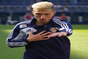 Ein junger Mann in einem blauen T-Shirt steht auf einem Fußballfeld und hat die Arme ausgestreckt und einen entschlossenen Gesichtsausdruck, der ihn als Flügelspieler ausweist.