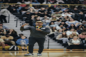 Ein Mann mit Mütze und Mikrofon auf einem Basketballfeld, dahinter Menschen auf einer Treppe und Taschen auf dem Boden.