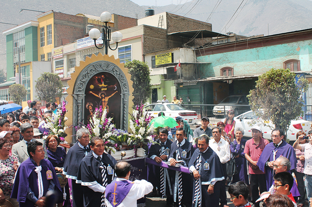 Menschen auf einer Stra├če vor der Stadt, mit einem Kruzifix und Blumenstr├Ąu├če im Vordergrund, fahrende Autos im Mittelgrund und Geb├Ąude, B├Ąume und Stra├čenlaternen im Hintergrund.