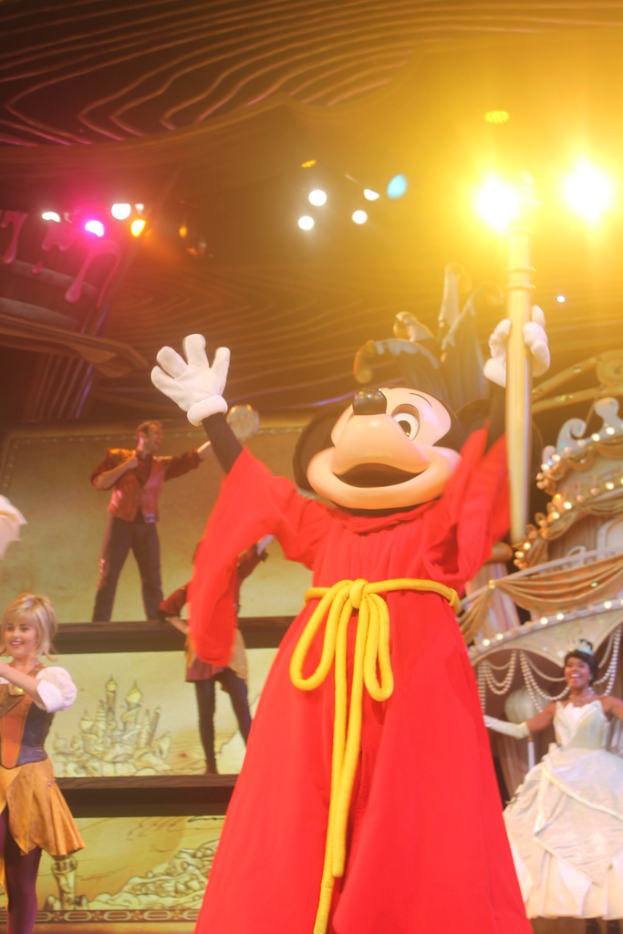 Eine Mickey-Maus-Puppe, die ein Objekt in der Vordergrund h├Ąlt, mit einer Gruppe von Menschen in verschiedenen Kost├╝men, die im Hintergrund gegen eine Wand und ein Dach mit Lichtern auftreten.
