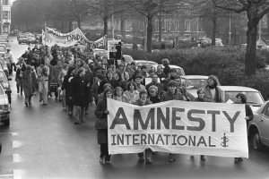 Eine Gruppe von Menschen marschiert mit einem Banner, auf dem 'Amnesty International' steht, eine Straße entlang, mit parkenden Autos auf beiden Seiten und Bäumen und Gebäuden im Hintergrund, dargestellt in Schwarz-Weiß.