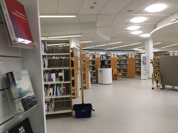 Ein geräumiger Bibliotheksinnenraum mit zahlreichen Bücherregalen, die mit Büchern gefüllt sind, einem Korb auf dem Boden, einem Giraffen-Spielzeug, Stühlen, Tischen, Säulen und Deckenleuchten.