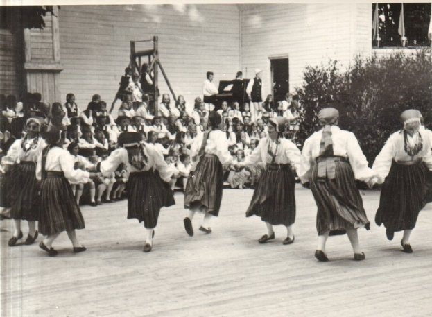 Eine Gruppe von Frauen, die vor einem gemischten Publikum aus sitzenden und stehenden Menschen tanzen, mit einem Gebäude, Pflanzen und einem hölzernen Objekt im Hintergrund, dargestellt in Schwarz-Weiß.