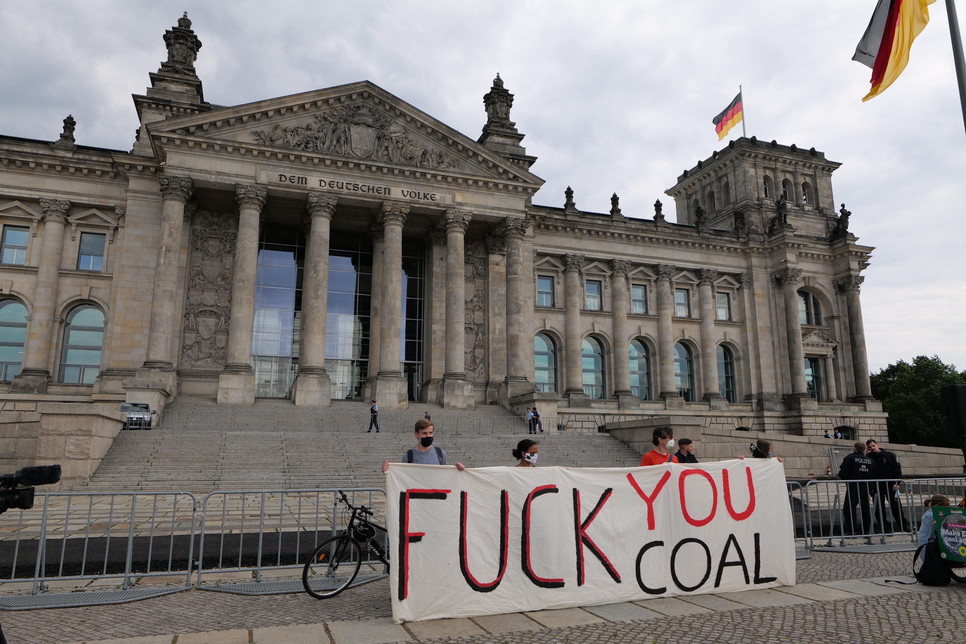Eine Gruppe von Menschen hält ein "Fuck You Coal"-Schild vor dem Reichstag in Berlin, mit Bäumen, einer Fahnenstange und einem bewölkten Himmel im Hintergrund.