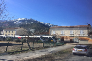 Ein Auto auf der linken Seite mit Zügen auf den Schienen vorne, Gebäude im Hintergrund, Bäume auf der rechten Seite vor einem Zaun, ein Berg und Himmel darüber.