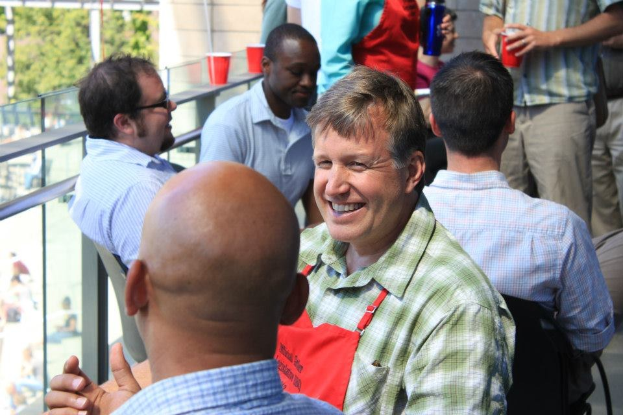 Ein Mann in einer Schürze, identifiziert als der Küchenchef der Universität Texas in Austin, spricht mit einem anderen Mann auf einem Balkon, während eine Gruppe von Menschen um ihn herum sitzt und steht; Bäume und ein Gebäude sind im Hintergrund zu sehen.