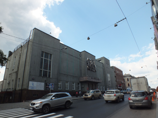 Eine Stadtstraße mit Autos, einer Zebrastreifen, Verkehrskegeln, Fußgängern, Gebäuden, Laternen, Strommasten mit Drähten, Bäumen und einer bewölkten Himmel.