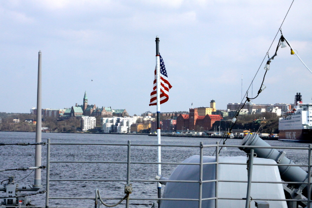 Eine Stadtansicht mit einem Schiff im Vordergrund, das eine Flagge zeigt, Booten auf dem Wasser, Gebäuden, Bäumen und Polen im Hintergrund, unter einem bewölkten Himmel.