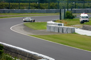 Ein Rennwagen fährt eine Rennstrecke entlang, im Hintergrund ist ein Polizeiwagen zu sehen, umgeben von Gras, Pflanzen, Bäumen und einem Fahrzeug auf der rechten Seite.