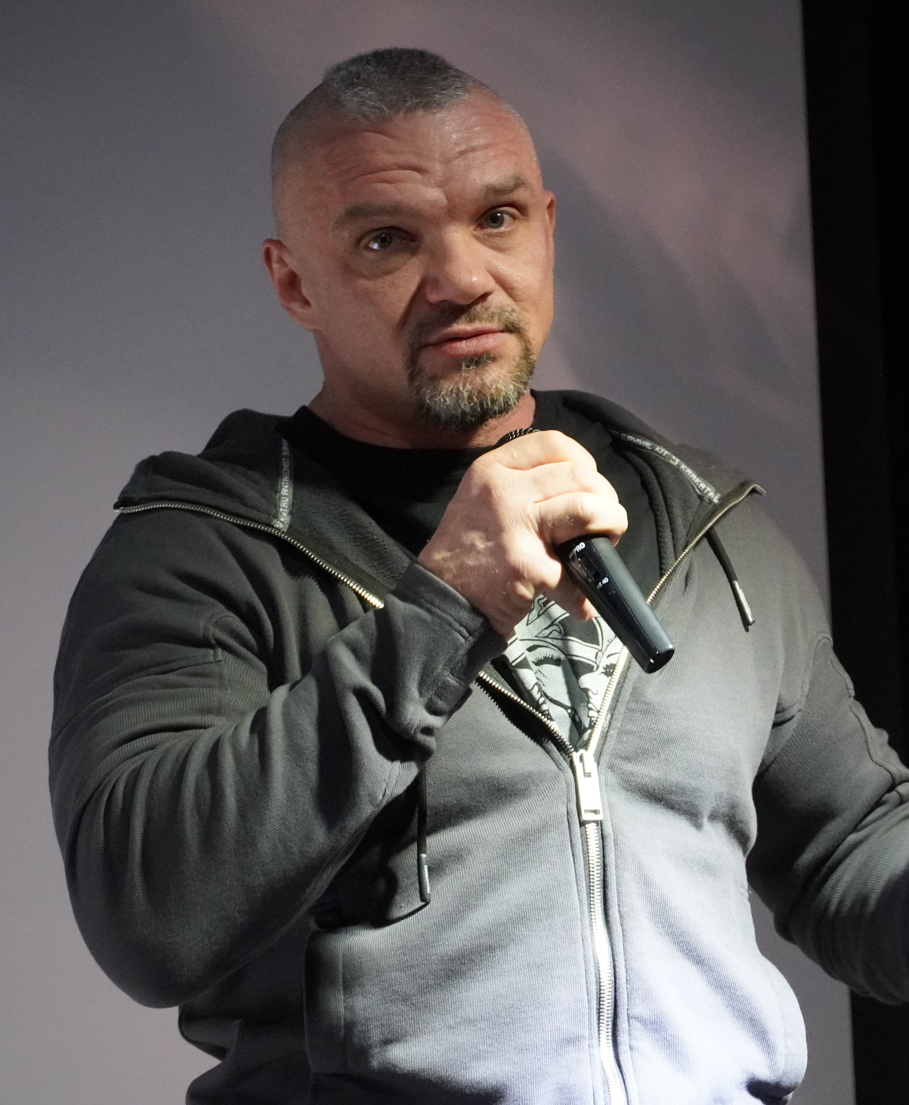 Ein Mann in einer schwarzen Kapuzenjacke und Anzug, der ein weißes Hemd trägt und das Haar zurückgekämmt hat, hält ein Mikrofon und steht selbstbewusst vor einer Wand.