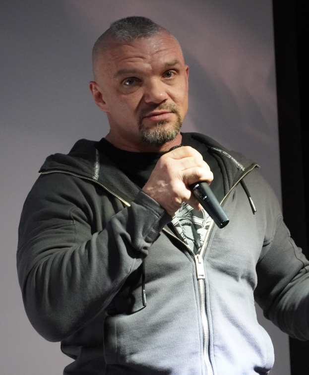 Ein Mann in einer schwarzen Kapuzenjacke und Anzug, der ein weißes Hemd trägt und das Haar zurückgekämmt hat, hält ein Mikrofon und steht selbstbewusst vor einer Wand.