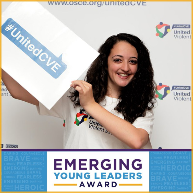 Eine strahlende Frau in einem weißen T-Shirt und Pferdeschwanz hält ein Schild mit der Aufschrift "Emerging Young Leaders Award" hoch und zeigt Freude und Stolz.