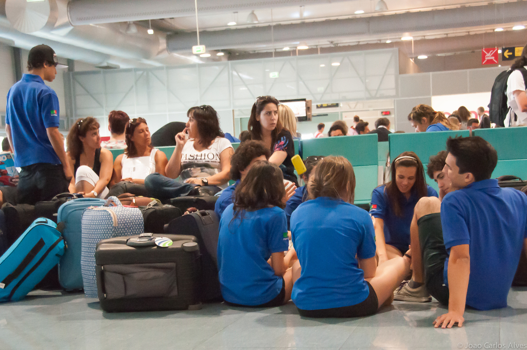 Ein belebtes Flughafeninnere mit vielen Menschen, die auf Flüge warten, Gepäckstücke links und Glastüren mit Hintergrundinformationen