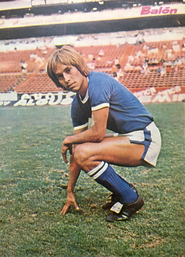 Ein Mann kniend auf einem Fussballfeld umgeben von Menschen auf Stühlen und Tafeln mit Text, trägt Schuhe.