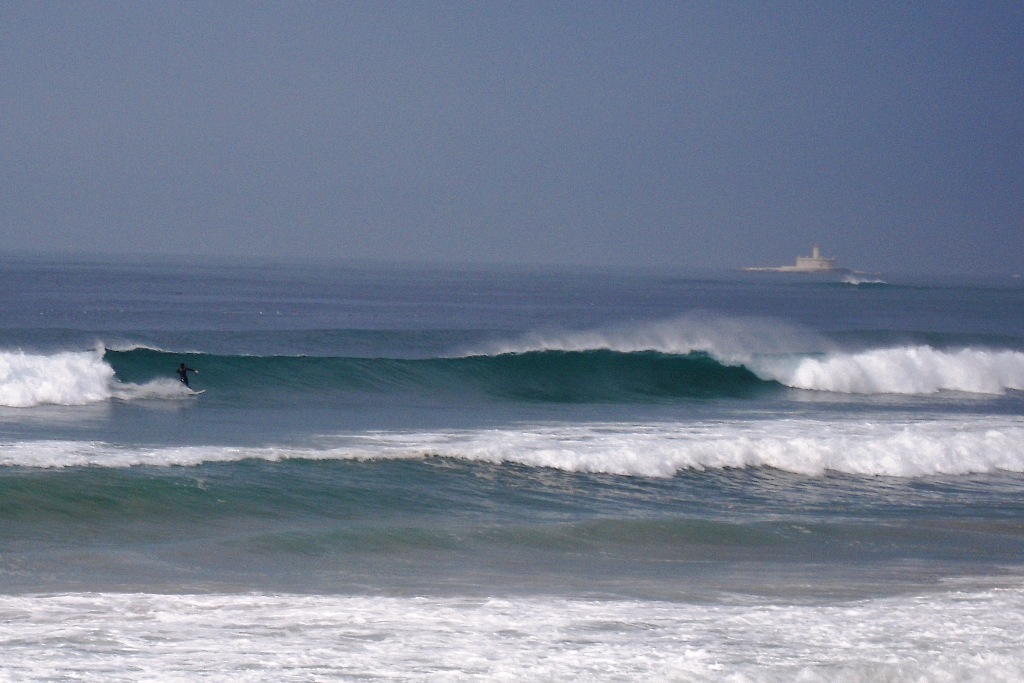 Eine Person surft auf einem Surfbrett im Wasser, mit einem Gebäude und dem Himmel im Hintergrund.