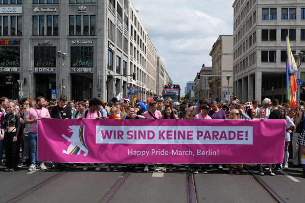 Eine Gruppe von Menschen, die eine Straße in Berlin, Deutschland, entlanggehen und ein pinkes Banner mit der Aufschrift "Happy Pride March" halten, mit Gebäuden, Laternenmasten und Verkehrszeichen an der Straße, unter einem bewölkten Himmel.