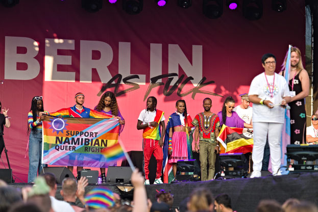 Gruppe von Menschen auf einer Bühne mit einem Banner, das "Berlin as Fuck" trägt, mit Lautsprechern, Mikrofonen und einer feiernden Menge unten.