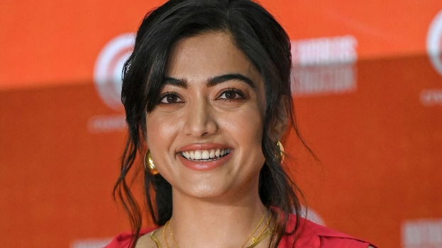 Frau in rotem Kleid mit elegantem Dutt und dezentem Make-up, die in die Kamera lächelt, vor einem gelben Banner mit der Aufschrift "Sonam Kapoors beste Schönheits Looks vom Cannes Film Festival."