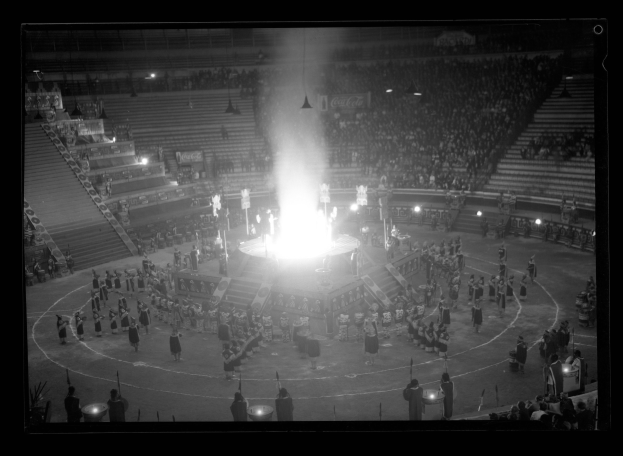 Ein Schwarz-Weiß-Foto einer Menschenmenge in einem Stadion, mit einer Bühne und einem brennenden Feuer in der Mitte, beleuchtet von Scheinwerfern und umgeben von Stufen und Geländern.