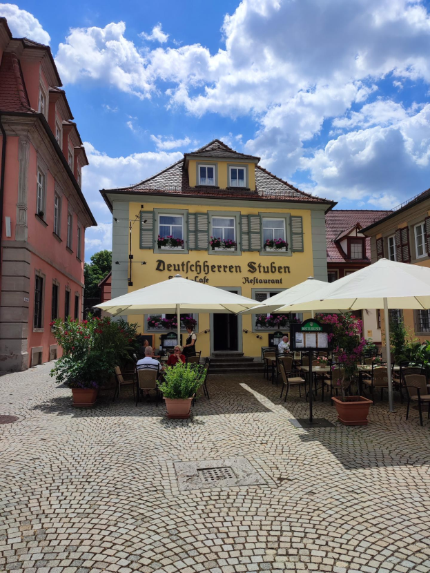 Ein Kopfsteinpflasterweg mit Tischen und Sonnenschirmen vor dem gelben Deutschen Stuben Restaurant in Baden-Württemberg, Deutschland, mit einigen Menschen, die sitzen und stehen, unter einem blauen und weißen Himmel.