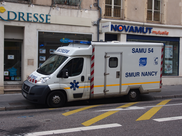 Ein Rettungswagen steht auf der Straßenseite vor einem Krankenhausgebäude mit Fenstern, Geländern und verschiedenen Schildern und Plakaten.