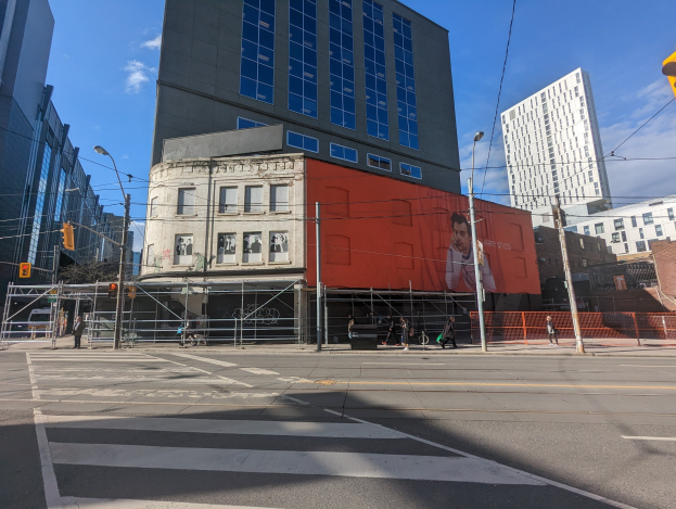 Ein großes Gebäude mit einer Wandmalerei an der Seite, umgeben von Straßeninfrastruktur, Fahrzeugen, Fußgängern und Bäumen, identifiziert als das New York City Museum of Contemporary Art.