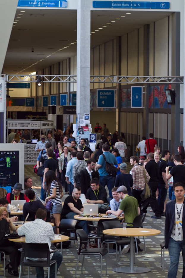 Ein belebter Flughafen-Lobby mit wartenden Menschen, einige stehen und andere arbeiten, Richtungsschilder an der rechten Wand und über, sowie von der Decke hängende Lichter.