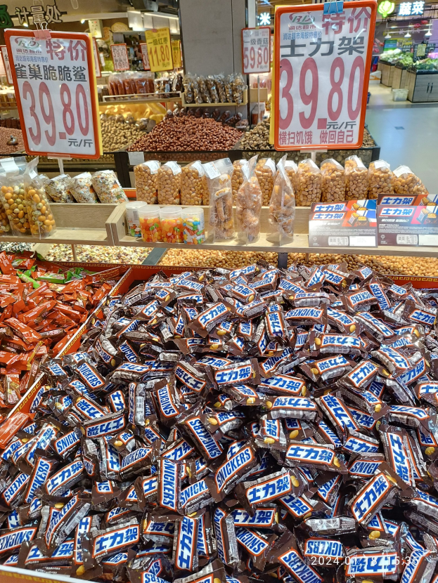 Vitrine mit verschiedenen Süßigkeiten, einschließlich Snickers-Riegeln, in einem Geschäft mit Lebensmitteln auf Regalen, beleuchteter Deckenbeleuchtung und Hintergrundwerbung.