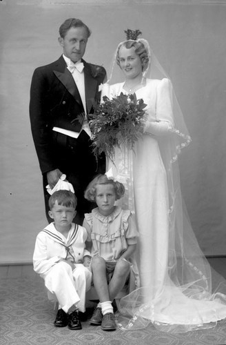 Schwarz-weiß-Foto einer Braut in einem weißen Kleid mit einem Strauß, einem grinsenden Bräutigam neben ihr und zwei Kindern, die vor ihnen auf dem Boden sitzen.