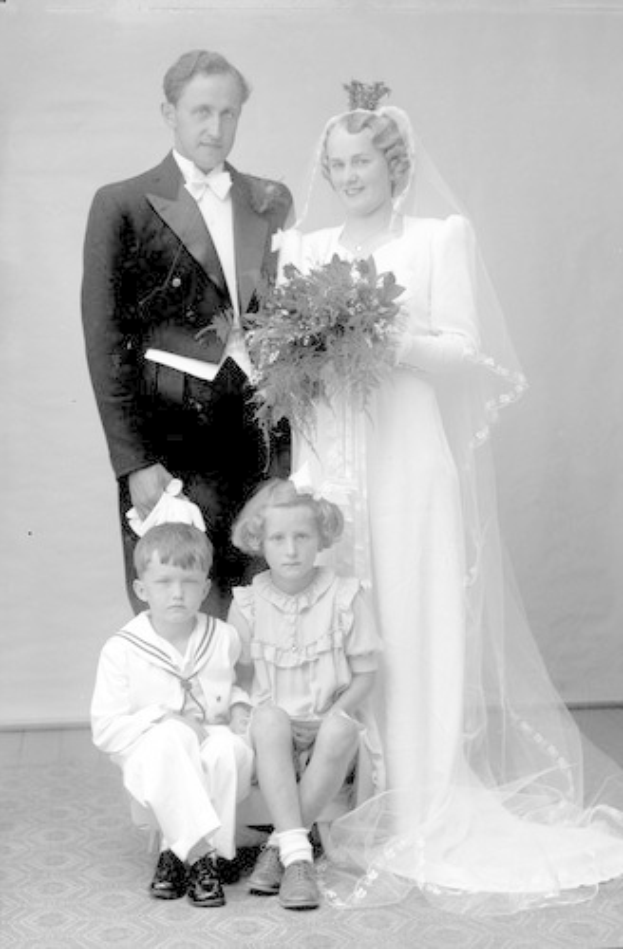 Schwarz-weiß-Foto einer Braut in einem weißen Kleid mit einem Strauß, einem grinsenden Bräutigam neben ihr und zwei Kindern, die vor ihnen auf dem Boden sitzen.