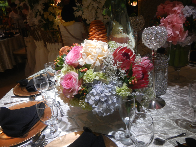 Formaler Esstisch mit Tellern, Besteck, Gläsern, Servietten und einem Blumenstrauß als Tischschmuck, umgeben von Stühlen und besetzten Tischen im Hintergrund.
