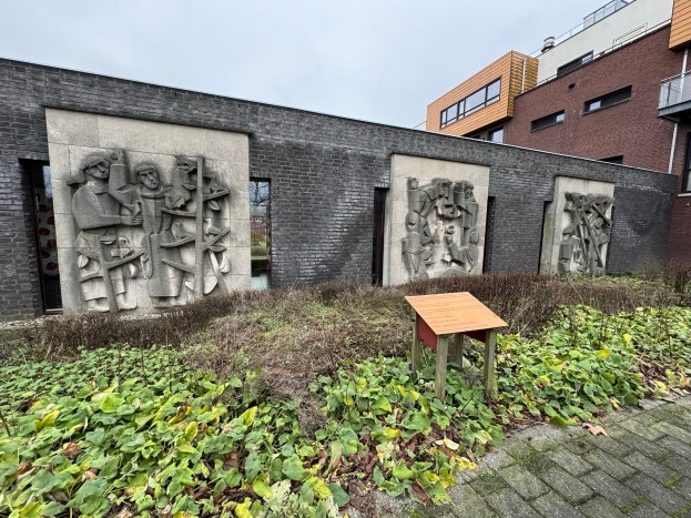 Ein Ziegelgebäude mit Skulpturen an seiner Seite, umgeben von Pflanzen und einer Texttafel, mit dem Himmel im Hintergrund, Teil des Museums für Moderne Kunst in Amsterdam.
