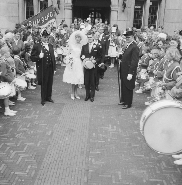 Schwarzes und weißes Foto einer Braut in einem weißen Kleid und Schleier und eines Bräutigams mit einem Blumenstrauß, der eine Straße entlang geht, gefolgt von einer Menge, von denen einige Musikinstrumente halten, vorbei an einem Gebäude mit einem Banner.