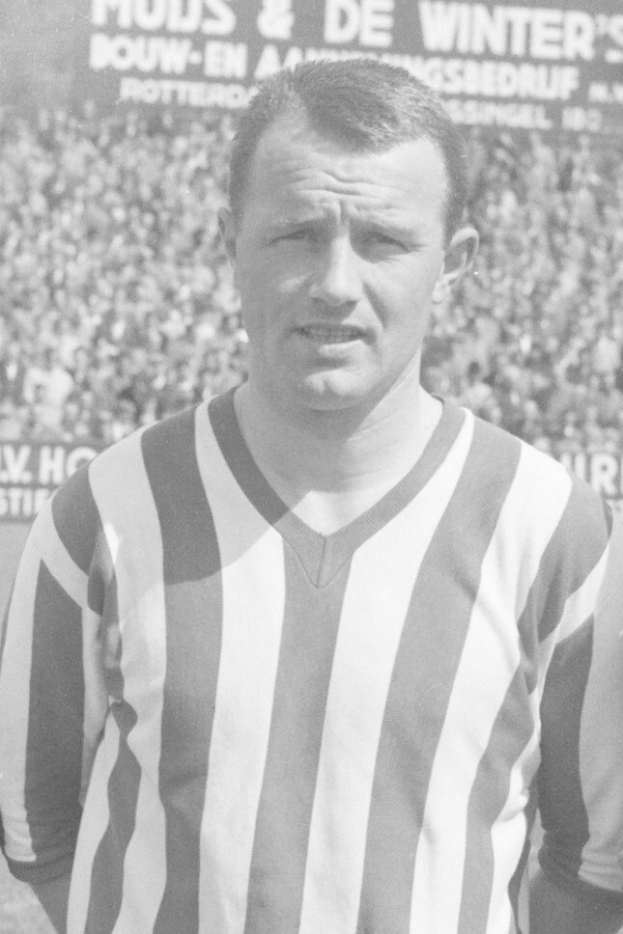 Ein Schwarz-Weiß-Foto eines Mannes in einem gestreiften Hemd, der vor einer Menge steht und ein Schild im Hintergrund hält, das als legendärer niederländischer Fußballspieler Bertrand identifiziert wird.
