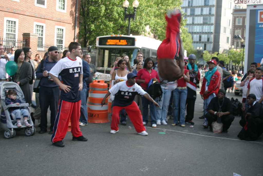 Eine Gruppe von Menschen, einige mit Mötzen, mit einem Kind im Kinderwagen, einem Ballon und einem Verkehrsobjekt im Vordergrund, einem Bus im Hintergrund und Gebäuden, Bäumen, einer Laterne und einem Geländer weiter hinten.