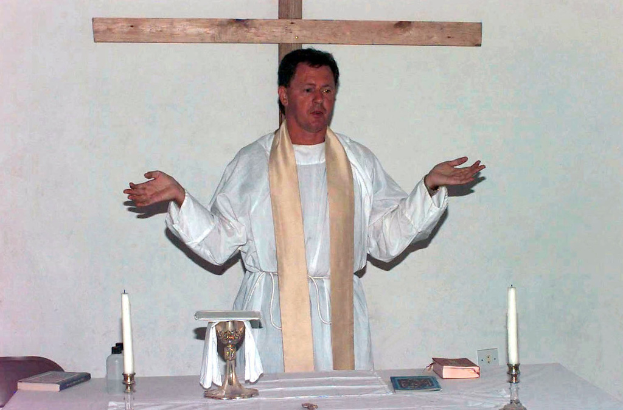 Ein Priester steht in der Mitte des Bildes mit einem Tisch vor ihm, der Kerzen und Bücher zeigt.