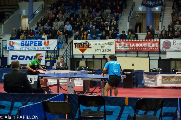 Zwei Männer spielen Tischtennis drinnen, mit mehreren Zuschauern, die stehen und sitzen, einigen leeren Stühlen und Werbebannern, mit Text in der linken unteren Ecke.