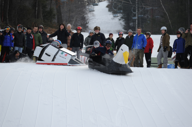 Menschen surfen auf dem Schnee mit vogel- und raketenförmigen Brettern, andere stehen dahinter, mit Seilbahnen und Bäumen im Hintergrund.
